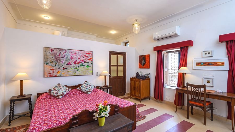 Room with bed, patterned bedding, furniture including desk, chair, tables, trunk, and cabinet, artwork, patterned rug, decorative ceiling lights, curtains, and patterned floor tiles - Tijara Fort-Palace - 19th Century, Alwar.