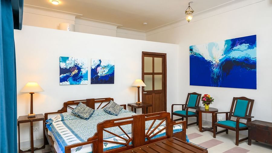 Room with bed, patterned pillows and runner, furniture including chairs, tables, and a bench, artwork, curtains, decorative ceiling lights, and patterned floor tiles - Tijara Fort-Palace - 19th Century, Alwar.