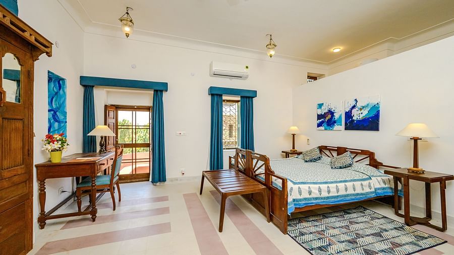 Room with bed, patterned pillows and runner, furniture including desk, chair, tables, and wardrobe, artwork, patterned rug, curtains, decorative ceiling lights, and patterned floor tiles - Tijara Fort-Palace - 19th Century, Alwar.