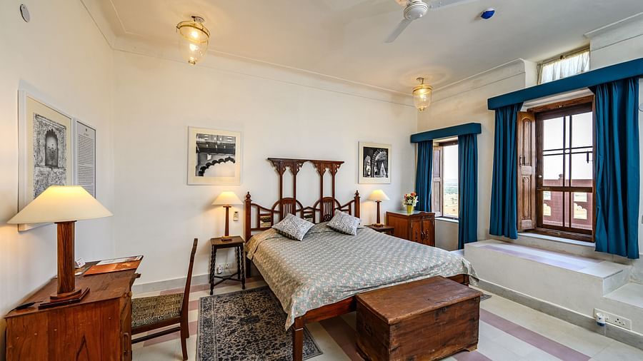 Room with bed, patterned pillows and bedding, furniture including desk, chair, tables, and a trunk, framed artwork, curtains, decorative ceiling lights, and patterned floor tiles - Tijara Fort-Palace - 19th Century, Alwar.