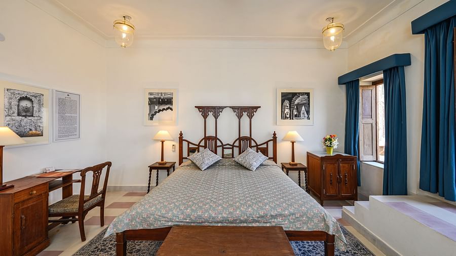 Room with bed, patterned pillows and bedding, furniture including desk, chair, tables, bench, and cabinet, framed artwork, curtains, decorative ceiling lights, and patterned floor tiles - Tijara Fort-Palace - 19th Century, Alwar.