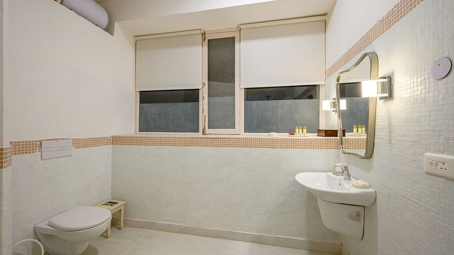 A bathroom with a toilet, a wall-mounted sink with a mirror above it, and windows with roller blinds at Tijara Fort-Palace - 19th Century, Alwar.