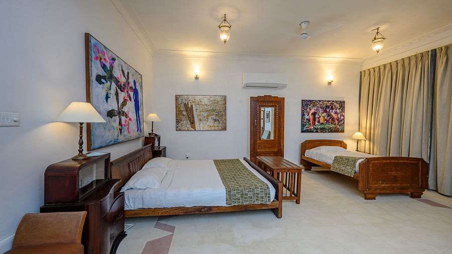 A spacious bedroom with two beds, wooden furniture including bedside tables with lamps and a wardrobe, artwork on the walls, and curtains on the windows at Tijara Fort-Palace - 19th Century, Alwar.
