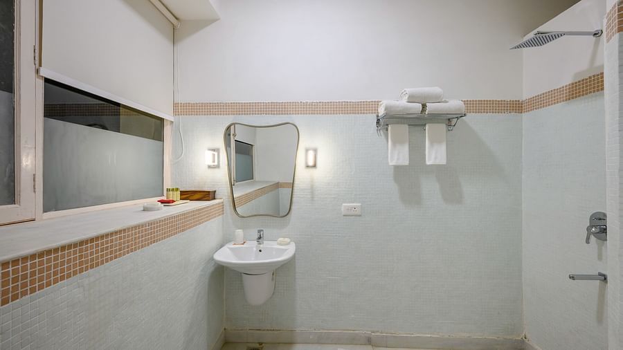 A bathroom featuring a wall-mounted sink with a mirror, a shower area with a glass partition, towel racks, and a window with a roller blind at Tijara Fort-Palace - 19th Century, Alwar