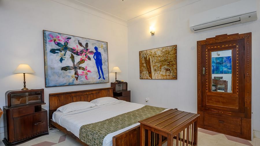 A bedroom with a bed, wooden bedside tables with lamps, artwork on the walls, and a wooden wardrobe with a mirrored door at Tijara Fort-Palace - 19th Century, Alwar.