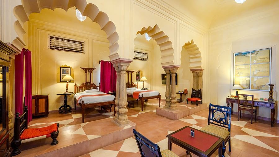 A room with two beds, two tables, chairs, and an arched entryway in Tijara Fort-Palace - 19th Century, Alwar.