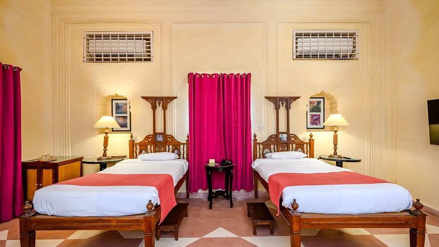 A bedroom with two beds, two side tables with lamps, and a small table in between in Tijara Fort-Palace - 19th Century, Alwar.