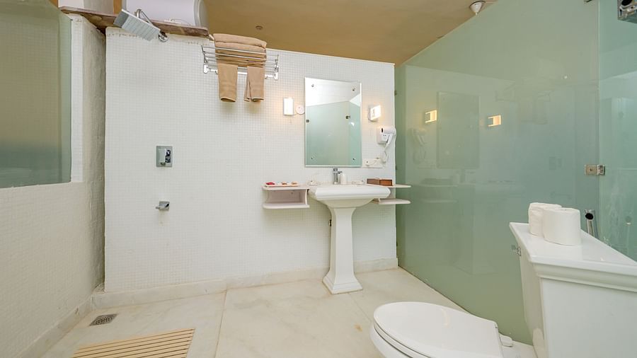 A bathroom with a toilet, a pedestal sink with a mirror above it, a shower area enclosed with frosted glass, and towel racks at Tijara Fort-Palace - 19th Century, Alwar.