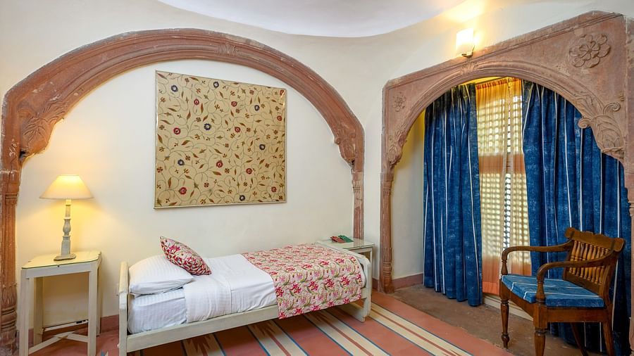 A bedroom with a single bed featuring patterned bedding, a bedside table with a lamp, an arched alcove with a tapestry, and another arched alcove with a chair and a window with curtains at Tijara Fort-Palace - 19th Century, Alwar.
