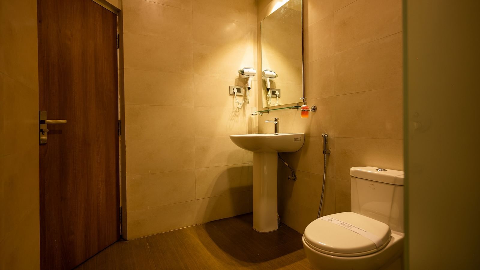 A well-lit modern bathroom with beige tiles, featuring a sink, wall-mounted mirror, toilet and amenities like hand soap and a hair dryer.