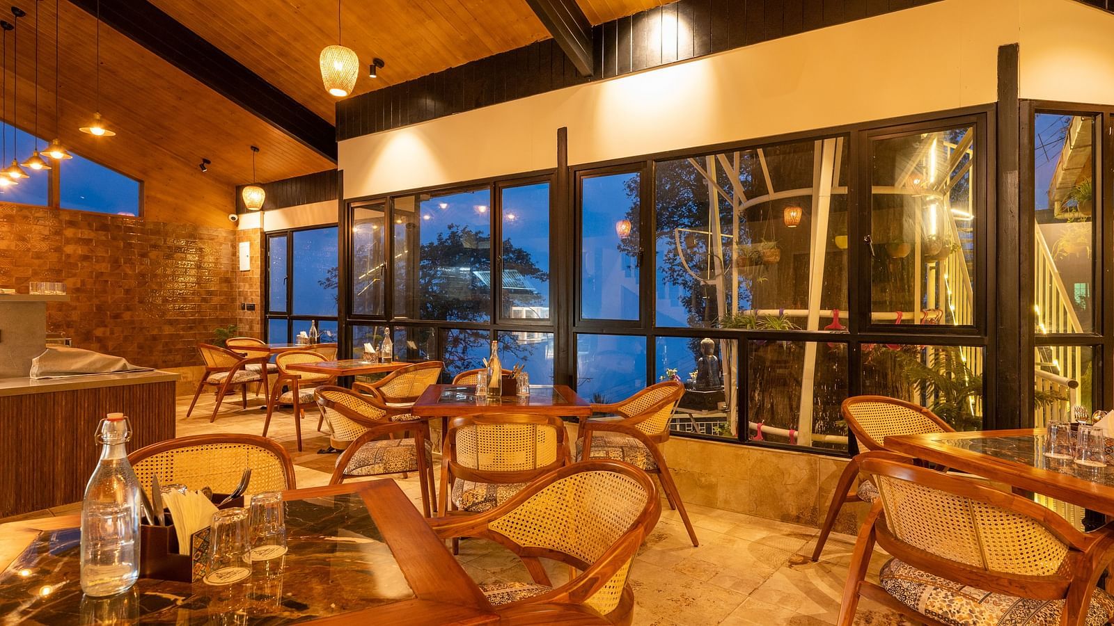 An elegant restaurant interior at dusk with large windows offering a view of the outside, warm lighting, and wooden furniture creating a cosy dining atmosphere- Arcadia Heritage Resort , Darjeeling