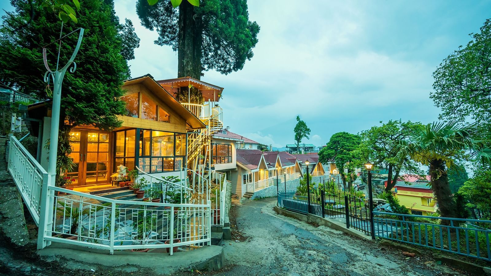 A wide view of cosy hillside establishment glowing warmly at twilight, with its wooden architecture and inviting lights reflecting a welcoming ambience amidst lush greenery- Arcadia Heritage resort, Darjeeling