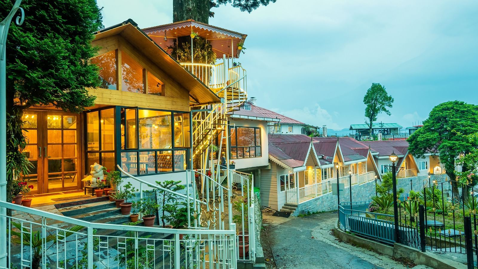 A close up view of a cosy hillside establishment glowing warmly at twilight, with its wooden architecture and inviting lights reflecting a welcoming ambience amidst lush greenery- Arcadia Heritage resort, Darjeeling