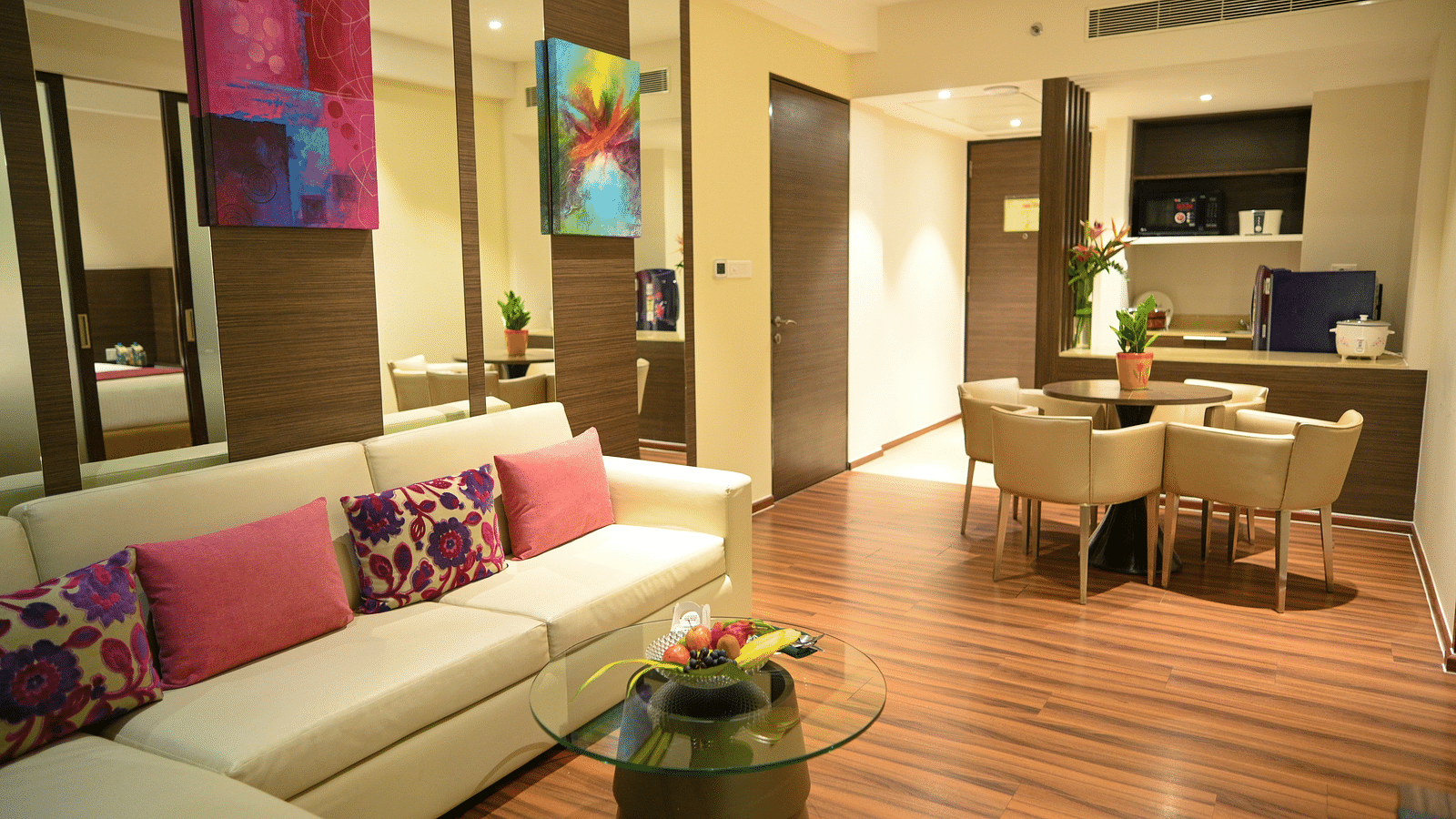 A well-lit living room with a sofa adorned with colourful pillows, a small coffee table, a dining table with chairs, and a compact kitchen.