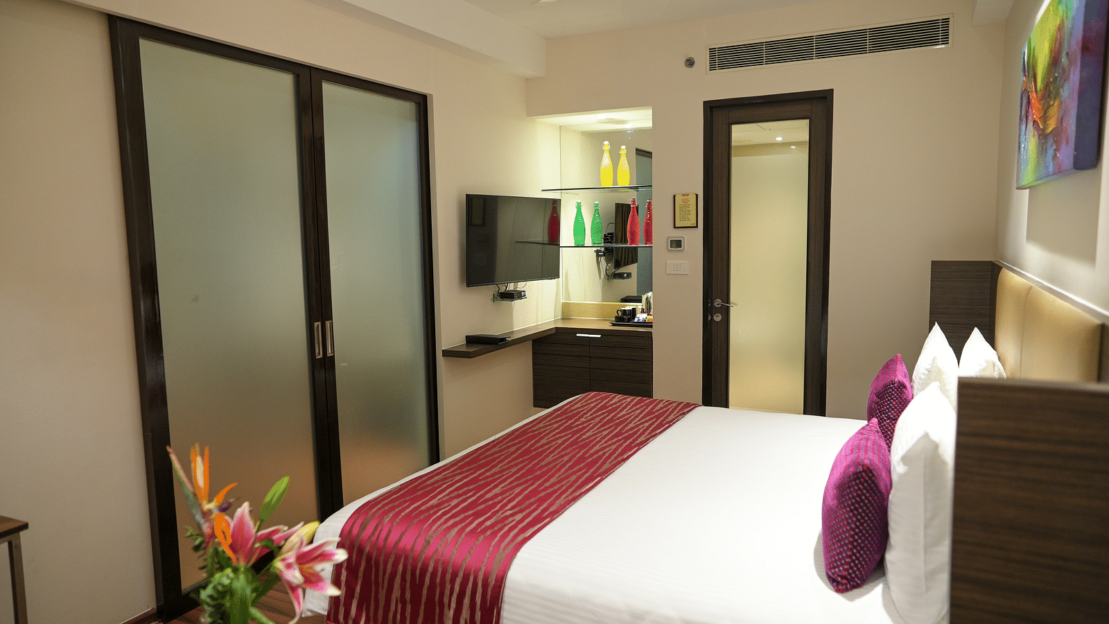 A well-lit hotel room with a king-sized bed, TV, and tea kettle. Doors lead to the bathroom and closet; a flower vase adorns a side table.