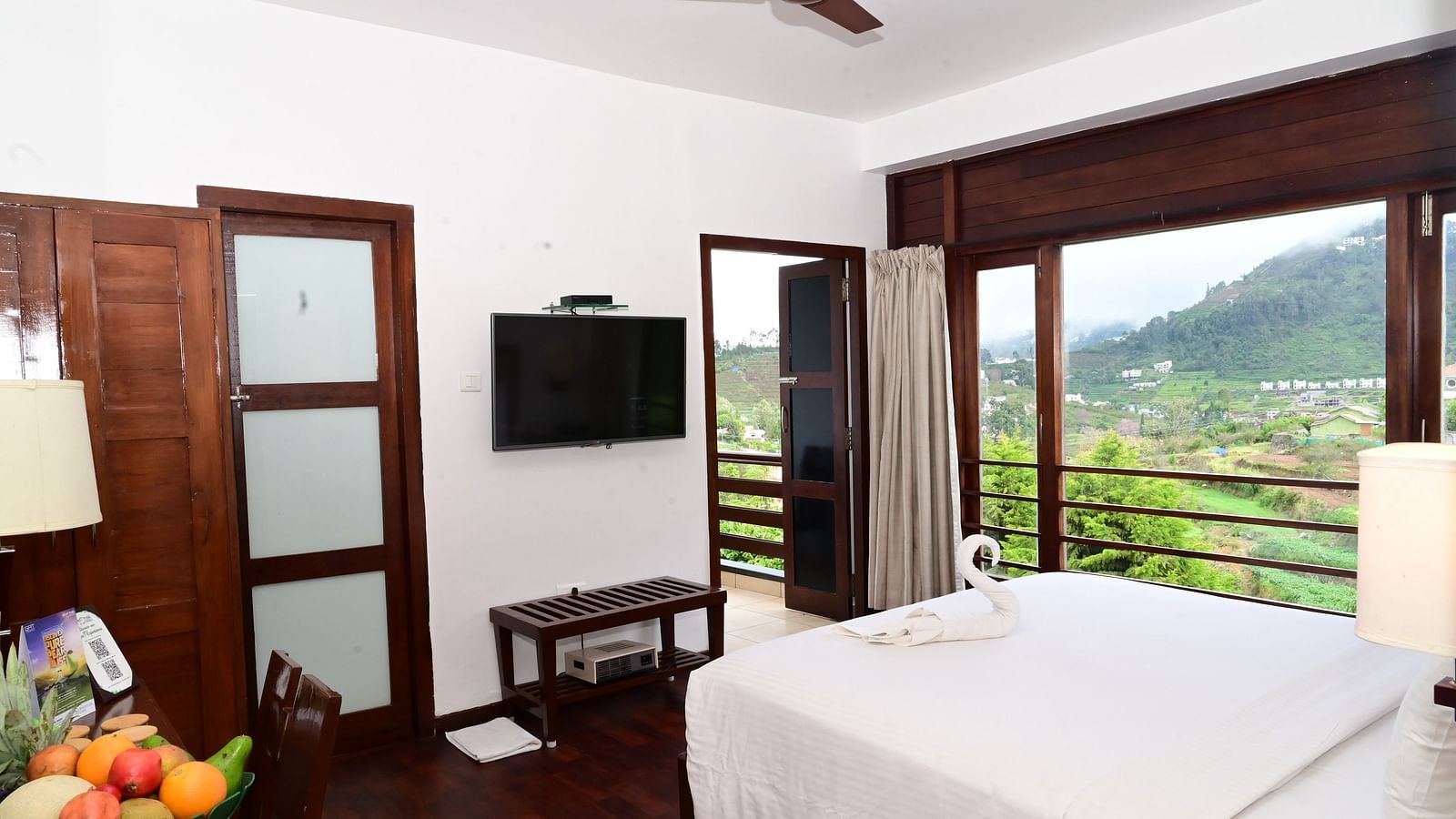 An image of a Rock Cabin Valley View with a king size bed, bed side table, lamps, flat screen TV, and an expansive window with curtains - GReaT trails Kodaikanal by GRT Hotels