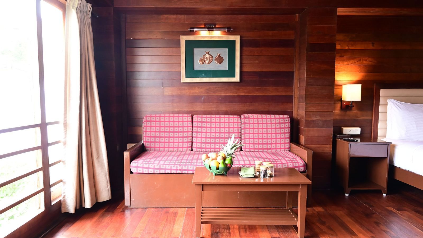 An image of a brightly lit room with a sofa, table, fruit basket, wall hangings, expansive window with curtains, and a bed - GReaT trails Kodaikanal by GRT Hotels