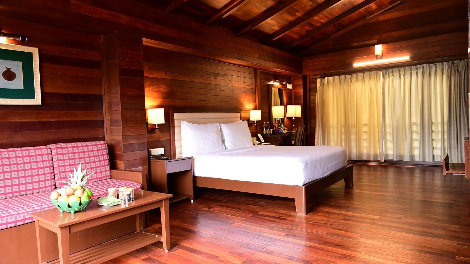 An image of a wood finish room with a king size bed, bed side table, lamps, sofa, table, and decorative items with bright lighting - GReaT trails Kodaikanal by GRT Hotels