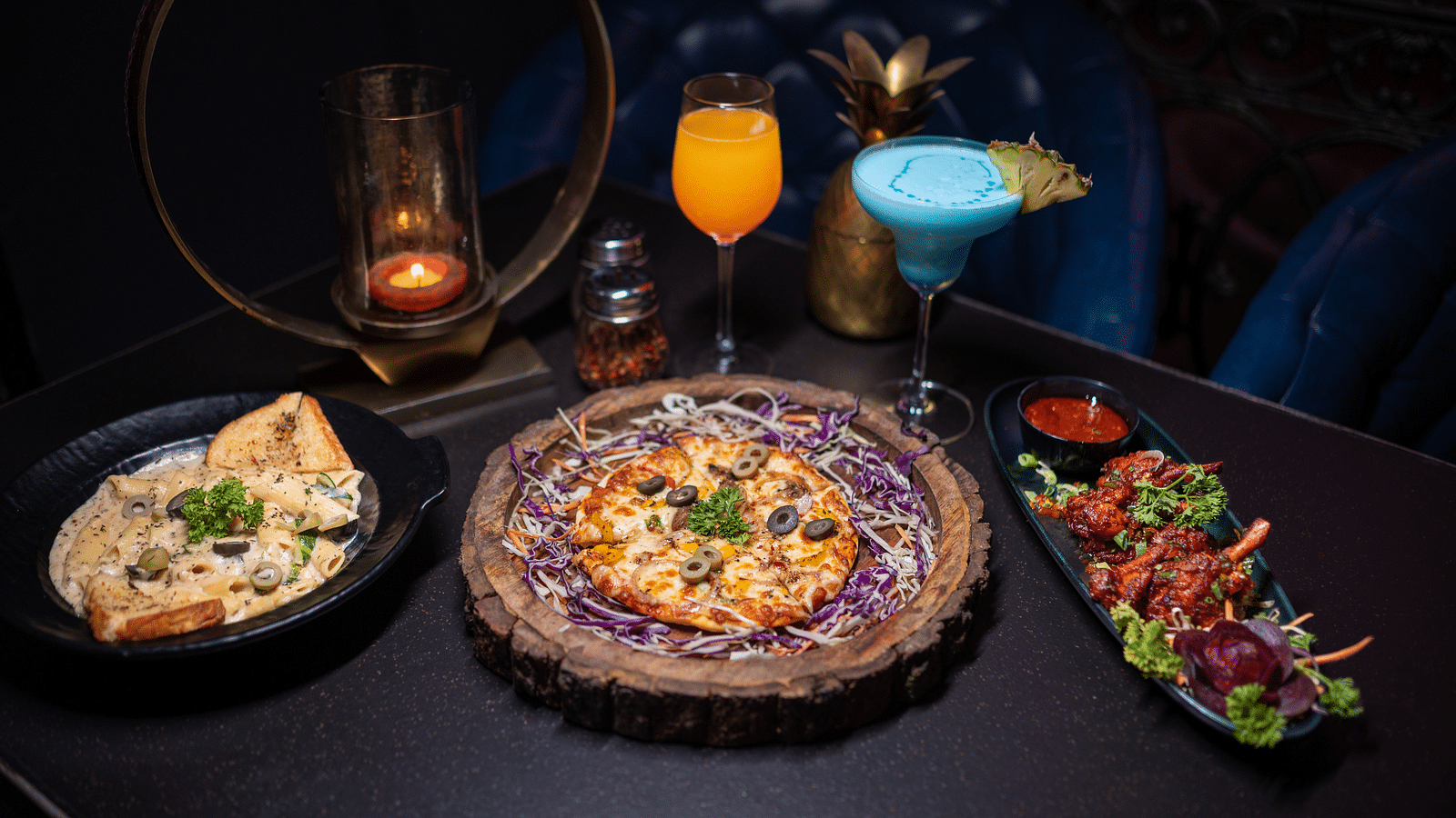 A top wide-angle view of a dining table featuring pizza, pasta, glasses of drinks, and starters, all beautifully arranged at Hotel Moti Palace.