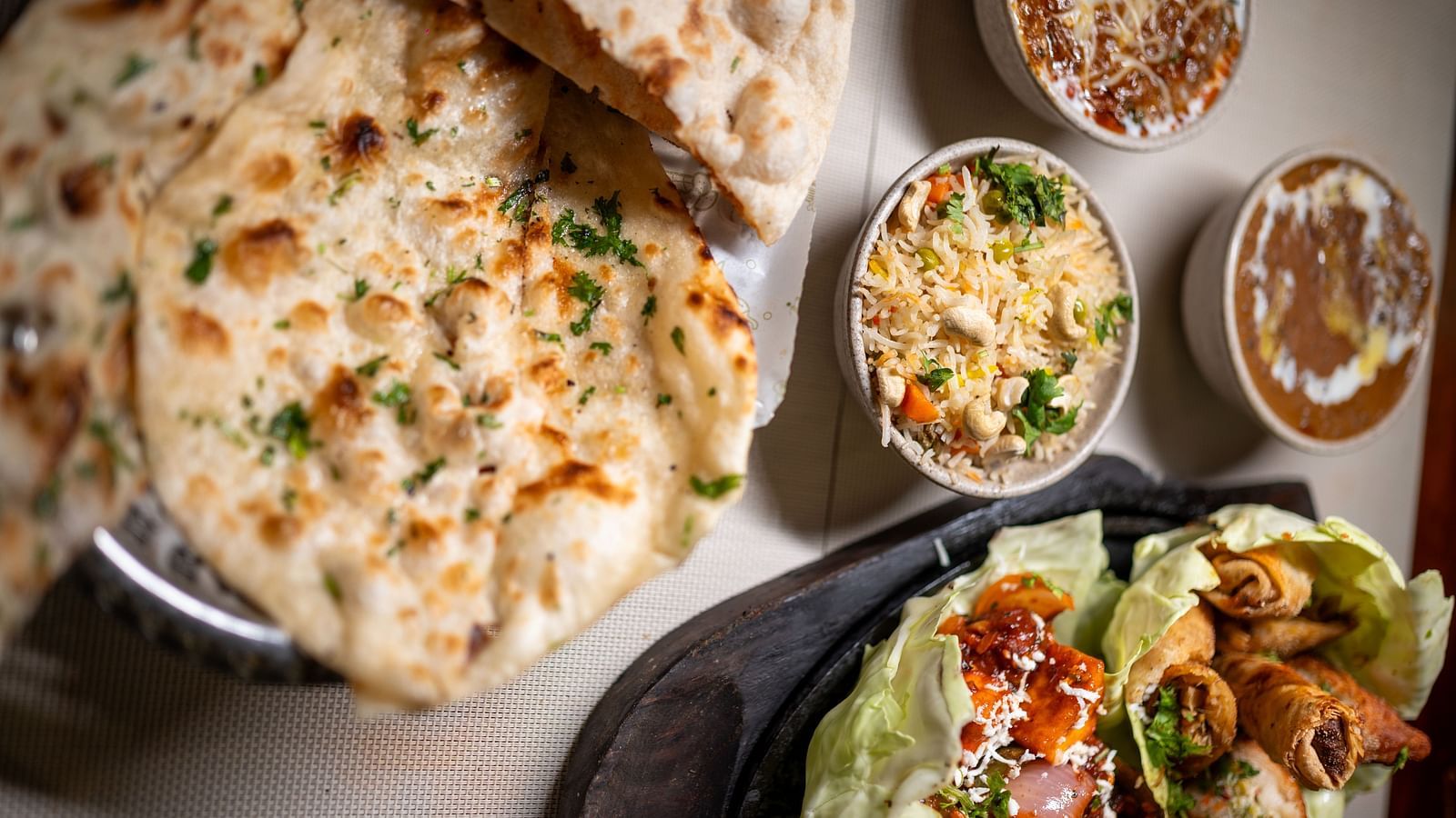 A beautifully arranged Indian meal on a table. The image features naan bread sprinkled with herbs, placed next to bowls of flavorful dishes including biryani, dal makhani, and curry. A sizzling plate of assorted appetizers like samosas and spring rolls is 