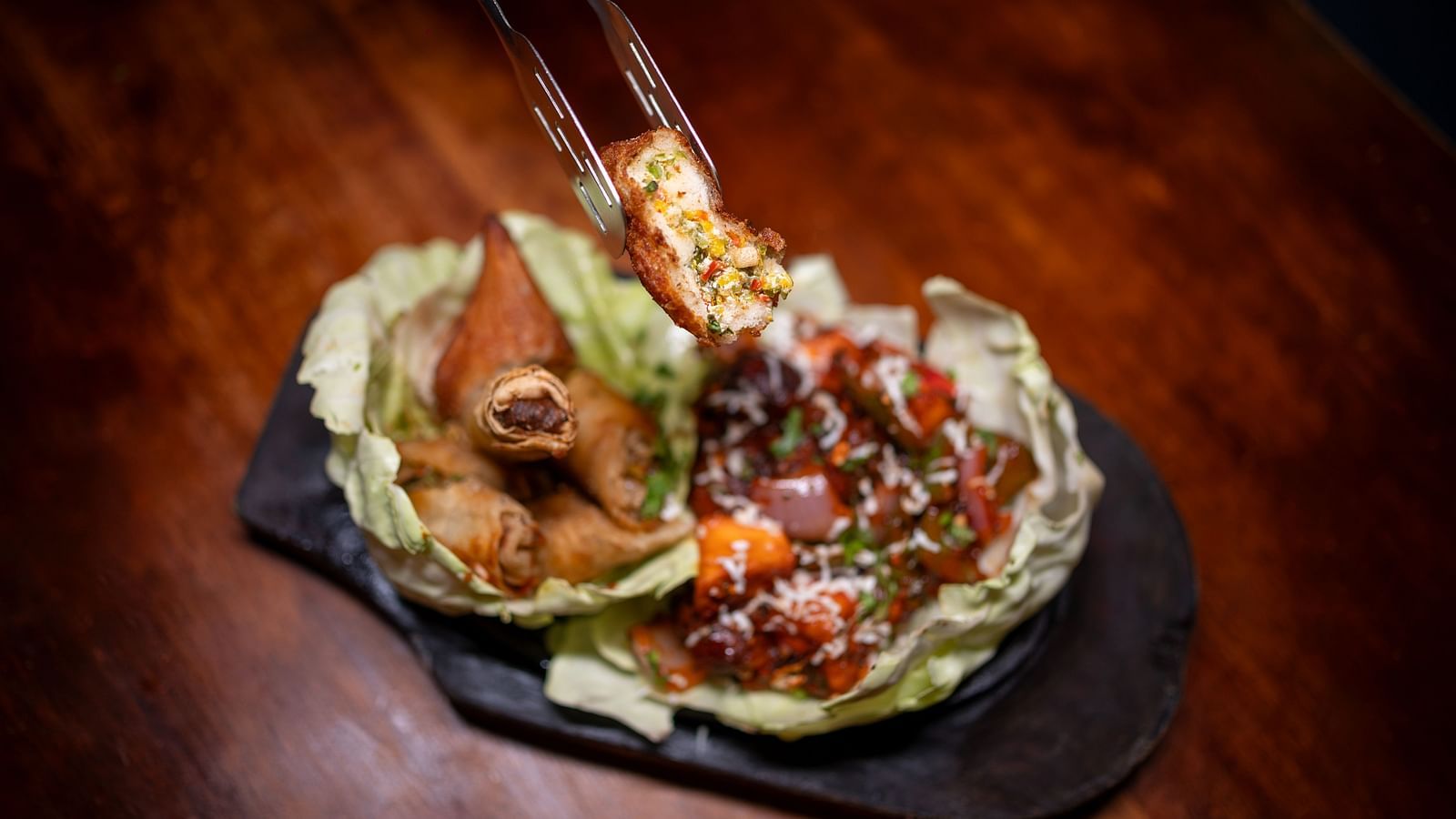  A close-up of tongs holding a crispy fried appetizer, possibly a stuffed spring roll or a paneer-filled snack. Below it, a plate displays other fried items alongside a vibrant vegetable stir-fry, all presented on cabbage leaves Hotel Moti palace ..