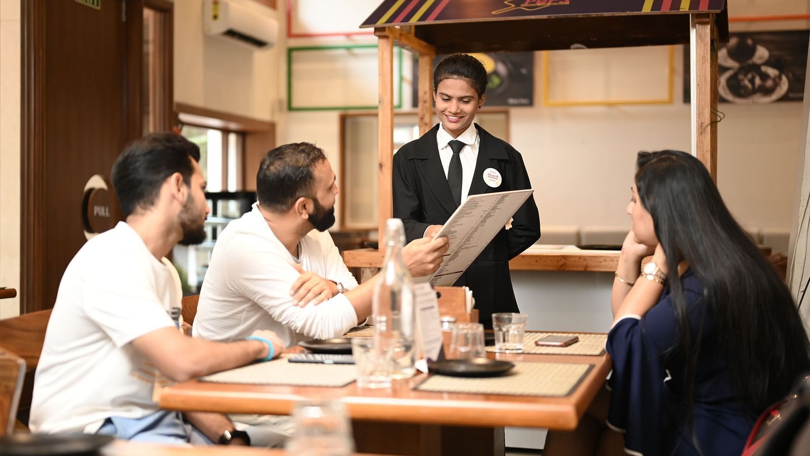 A hospitality trainee interacting with customers at a restaurant, holding a menu - Kamats India