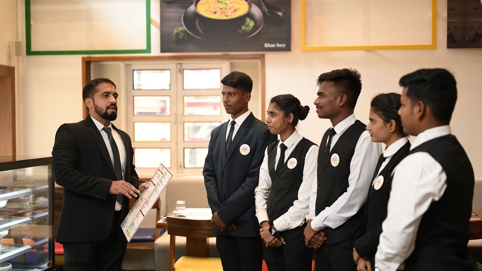 A manager in a suit explaining the menu to restaurant staff - Kamats India