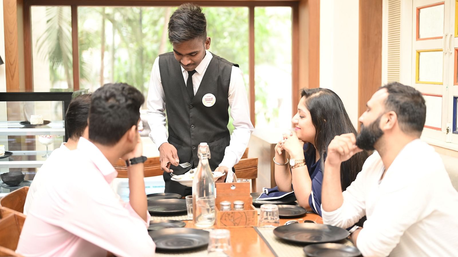 A waiter in a formal uniform serving food to guests in a restaurant - Kamats India