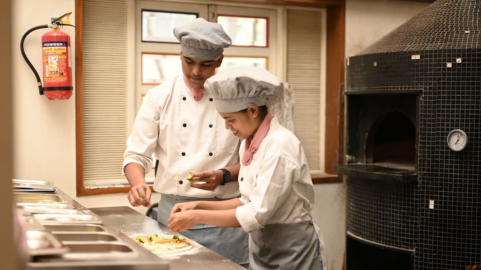 Two chefs garnishing a pizza before baking near a wood-fired oven - Kamats India