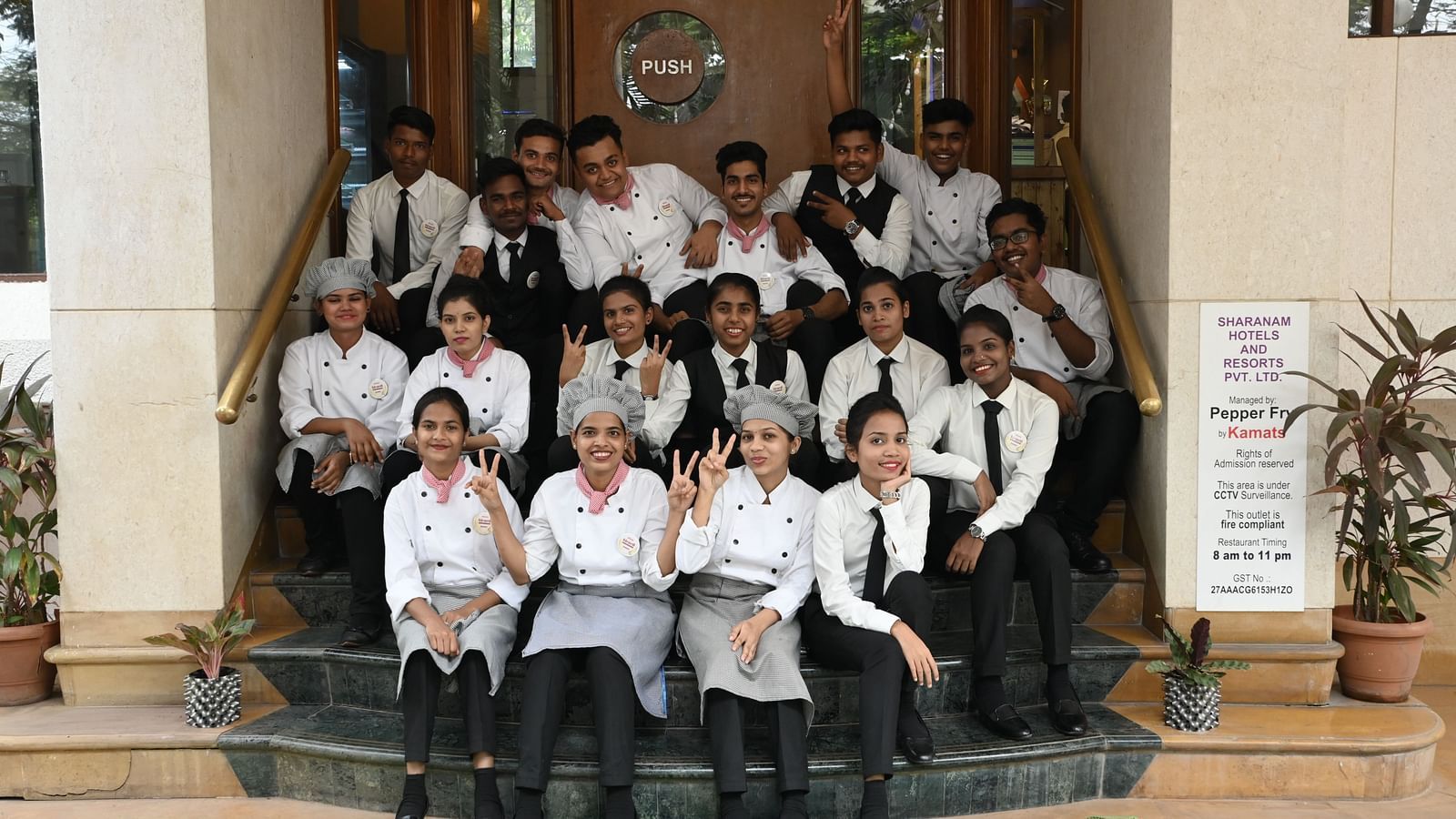 Hospitality and culinary trainees sitting on steps outside a hotel entrance, smiling and making peace signs - Kamats India