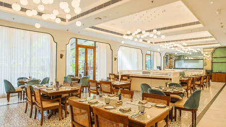 Bright restaurant dining room featuring wooden dining tables set with place settings, cane-back and teal upholstered chairs, modern cluster globe lights, and large sheer-curtained windows at Bhagirathi by Atishay