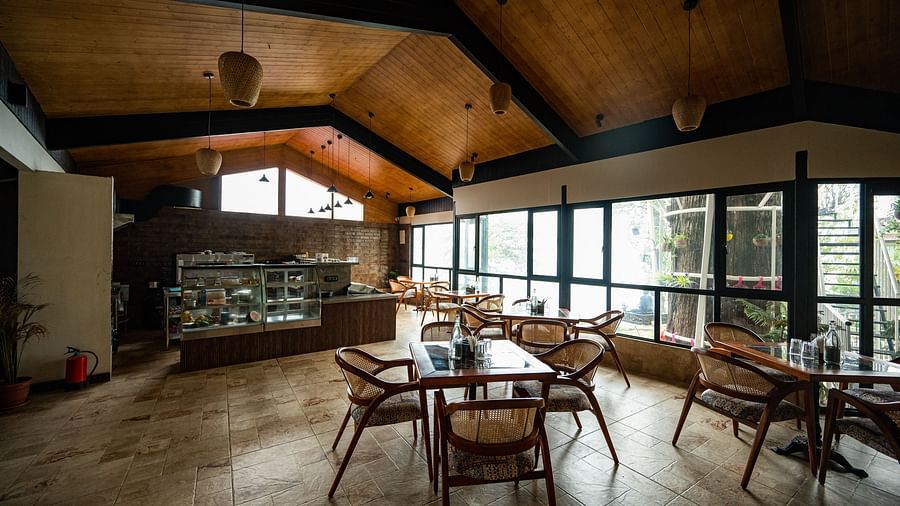 A spacious cafe interior with a high ceiling, featuring expansive windows, wooden furniture, and a counter displaying an array of treats, all bathed in the soft, natural light filtering through- Arcadia Heritage Resort, Darjeeling 