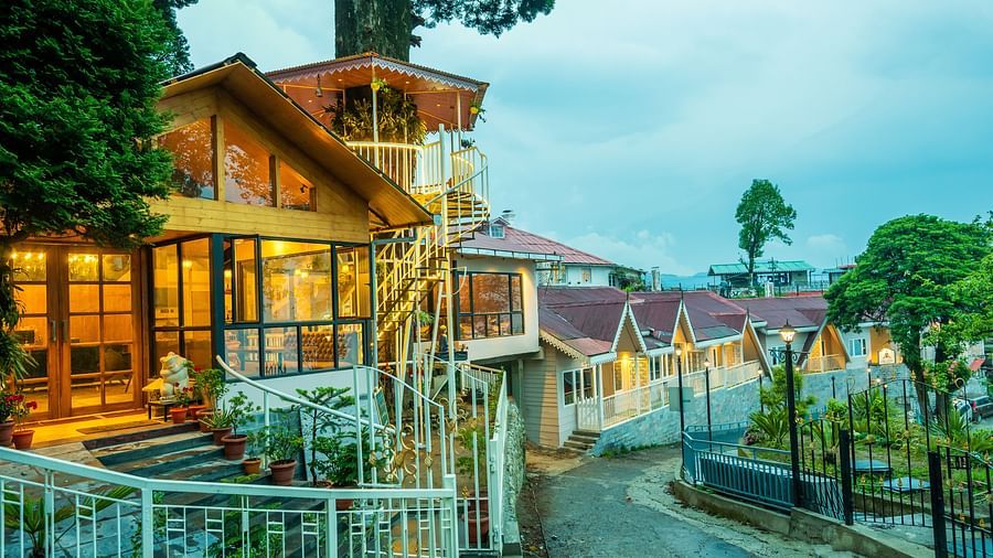 A close up view of a cosy hillside establishment glowing warmly at twilight, with its wooden architecture and inviting lights reflecting a welcoming ambience amidst lush greenery- Arcadia Heritage resort, Darjeeling