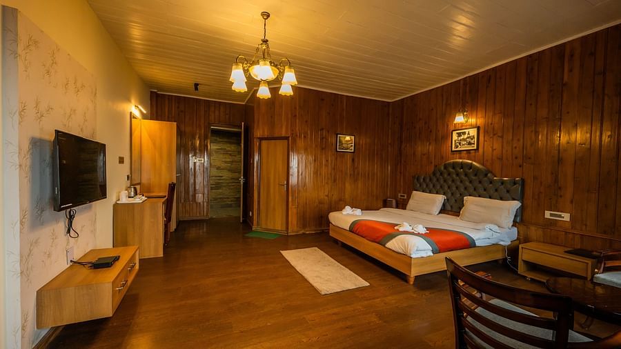 A side angle view of A spacious room with wooden furnished  comfy bed and the have bed side table and chair and Flat screen TV at Arcadia Heritage Resort, Darjeeling