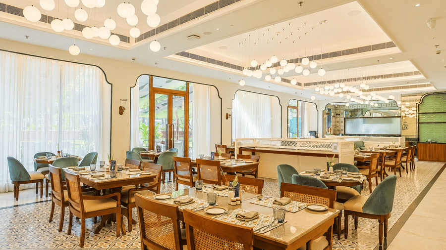 Bright restaurant dining room featuring wooden dining tables set with place settings, cane-back and teal upholstered chairs, modern cluster globe lights, and large sheer-curtained windows at Bhagirathi by Atishay