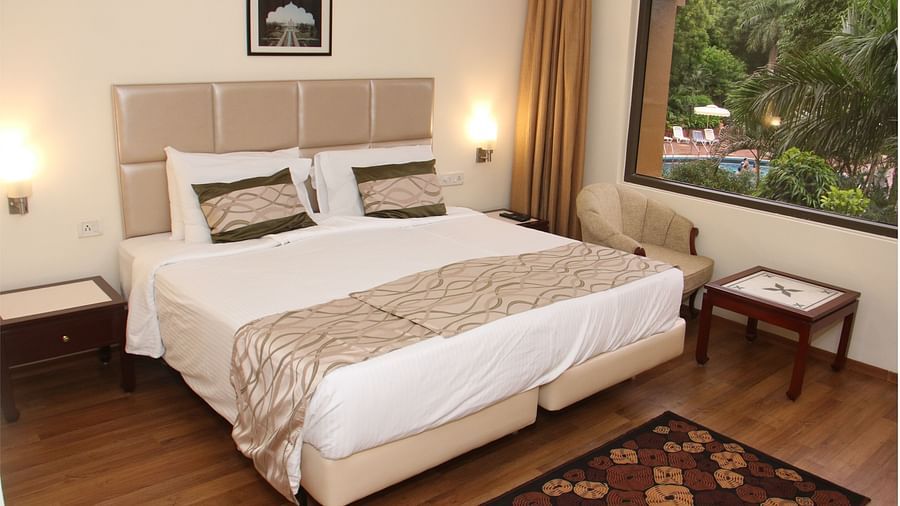 A room with a large bed, beige headboard, wooden flooring, bedside tables, and a window overlooking greenery and a swimming pool - Hotel Clarks Shiraz, Agra
