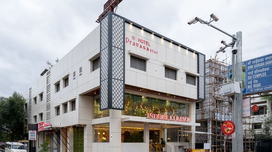 The exterior of Pravasa Stay, Shirdi is a three-storey building with large storefront windows at the ground level.