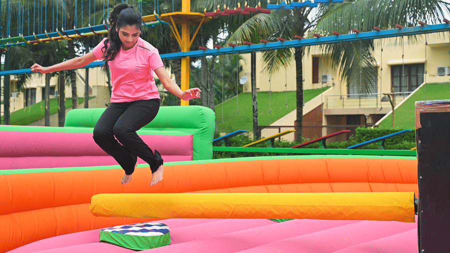 A girl jumping on colourful inflatable trampoline structures in an outdoor play area surrounded by trees at Vedic Village Spa Resort.
