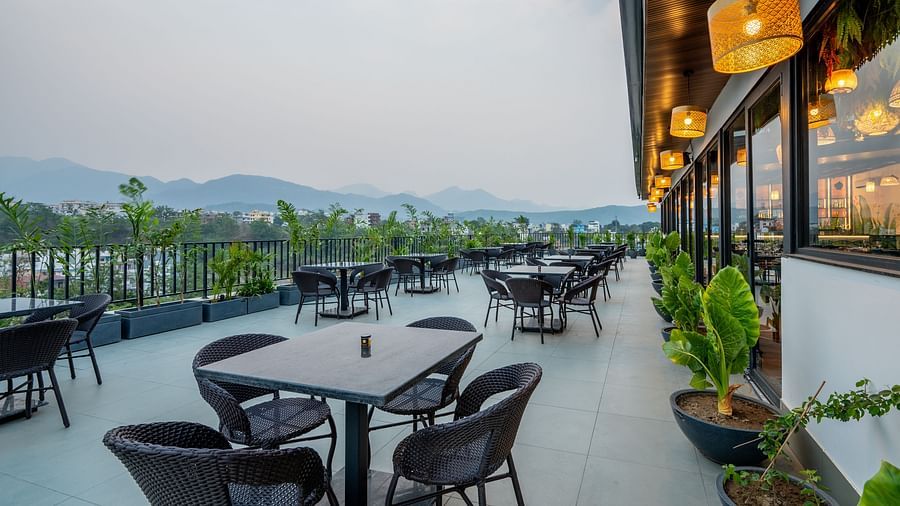 Side shot of an outdoor seating area at Vishranti Hotel, Dehradun with a seating area features multiple tables with chairs, and plants are visible in the background.