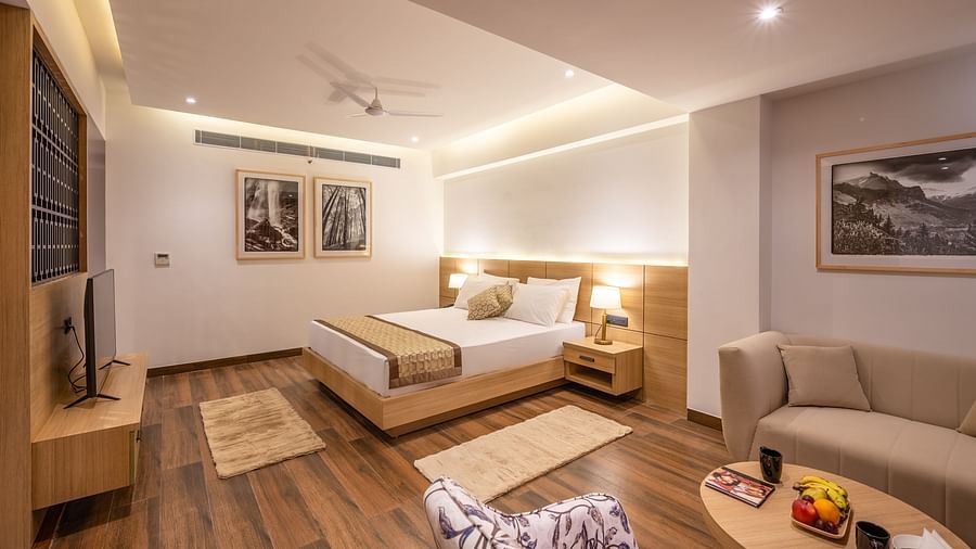 Side shot of Suite at Vishranti Hotel, Dehradun featuring a king bed with side tables, a sofa, a TV, large window with curtains and bed lamps. 