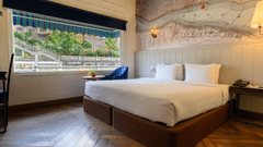 A modern hotel room featuring a large bed and a detailed map-like mural on the wall. A window with striped blinds overlooks lush greenery.