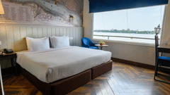 A stylish hotel bedroom featuring a bed with white bedding, a blue armchair, and a large window overlooking the water.