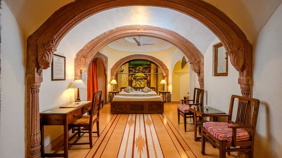 A hallway with a bed visible through an arched doorway, a desk, and seating at Tijara Fort-Palace - 19th Century, Alwar.