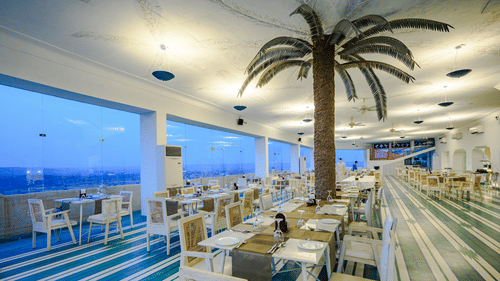 A dining area with multiple tables and chairs is visible inside Tijara Fort-Palace - 19th Century, Alwar, with large windows showing an outdoor view.