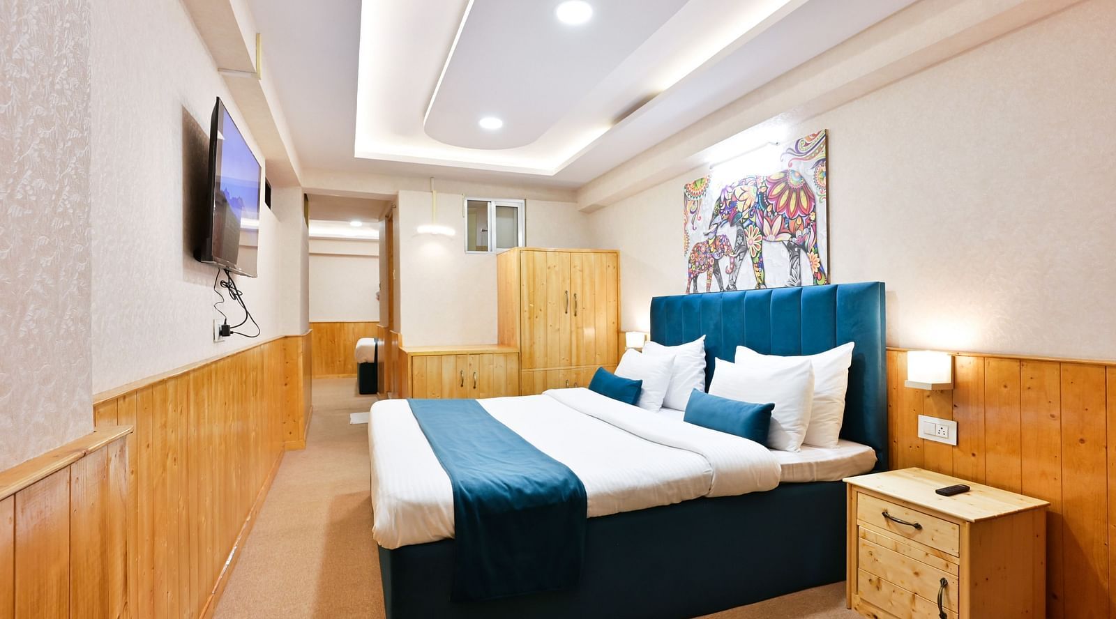Family room with a king size bed, wooden cupboards, wooden bedside table with bright lighting, and a wall-mounted television at Avianna Seven Seas, Darjeeling1