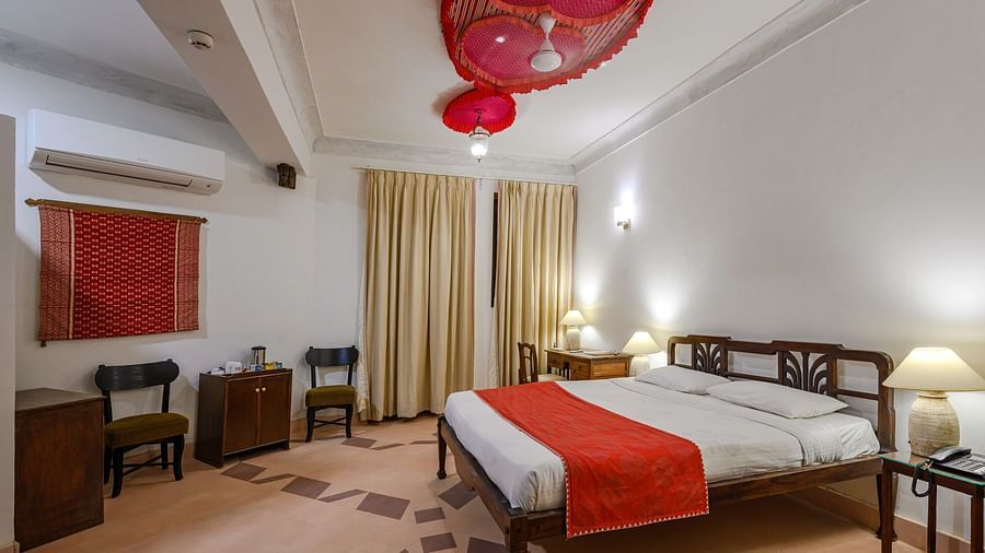 Tijara Fort-Palace - 19th Century, Alwar: Bedroom with a bed, bedside tables with lamps, a desk with a chair, two other chairs, and decorative ceiling elements.