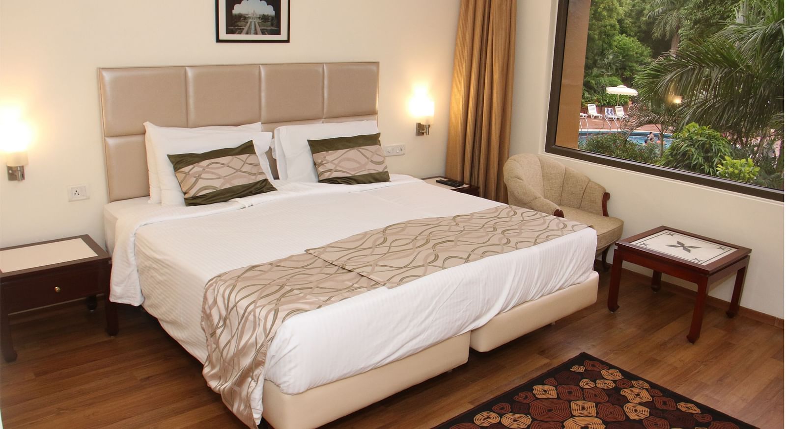 A room with a large bed, beige headboard, wooden flooring, bedside tables, and a window overlooking greenery and a swimming pool - Hotel Clarks Shiraz, Agra