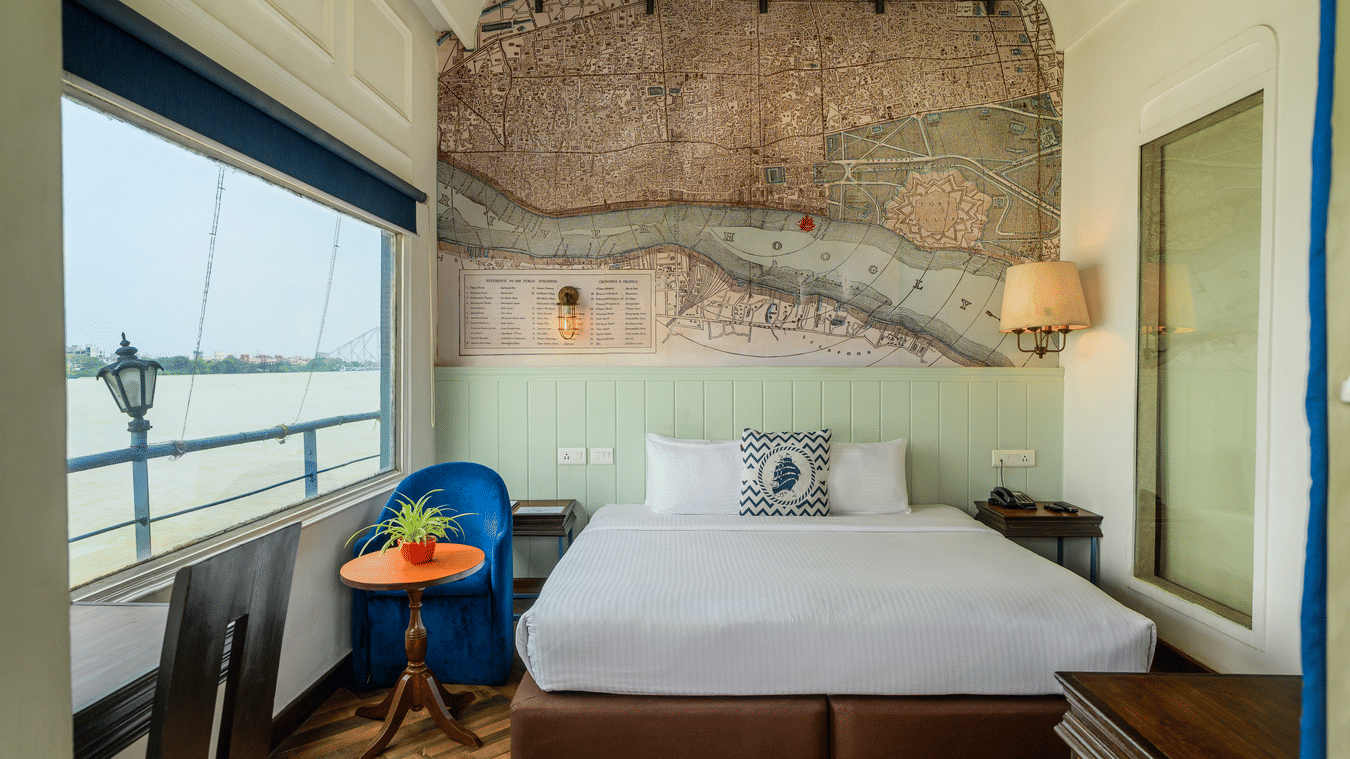 A hotel bedroom with a large bed, blue armchair, and a window with a view of the water and a lamp post. A vintage map mural is on the wall.