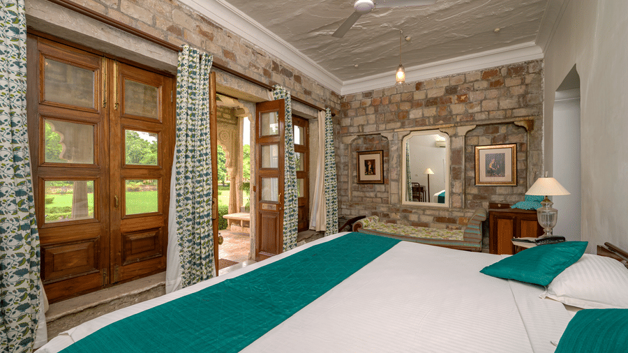Spacious heritage bedroom featuring carved wooden doors, stone walls, patterned curtains, elegant furnishings, and a large bed overlooking lush gardens at Deo Bagh, Gwalior