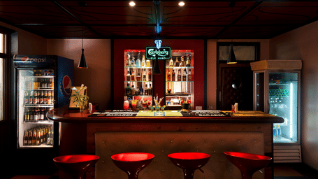 An indoor bar area, featuring a wooden counter lined with red stools, display fridges, and liquor bottles at Amantra Shilpi Resort, Udaipur.